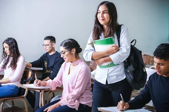 Classroom full of students with a female student standing up holding with books smiling.